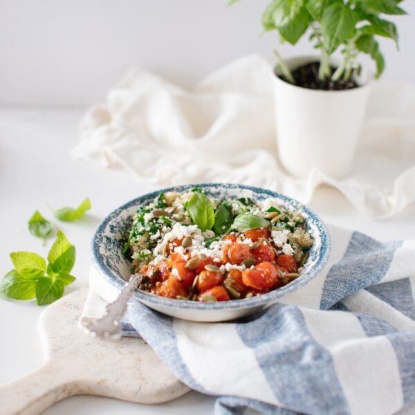 Bowl filled with quinoa and spinach and topped with roasted tomatoes and feta, with a basil plant in the background.