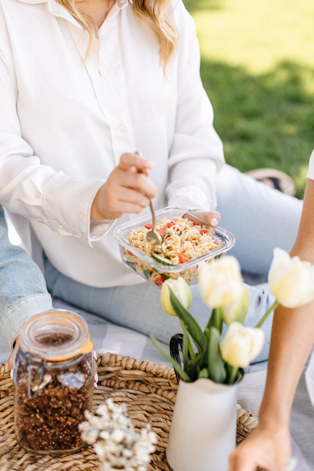 Girl in white blouse holding glass bowl of orzo salad on a picnic blanket with some flowers on it.