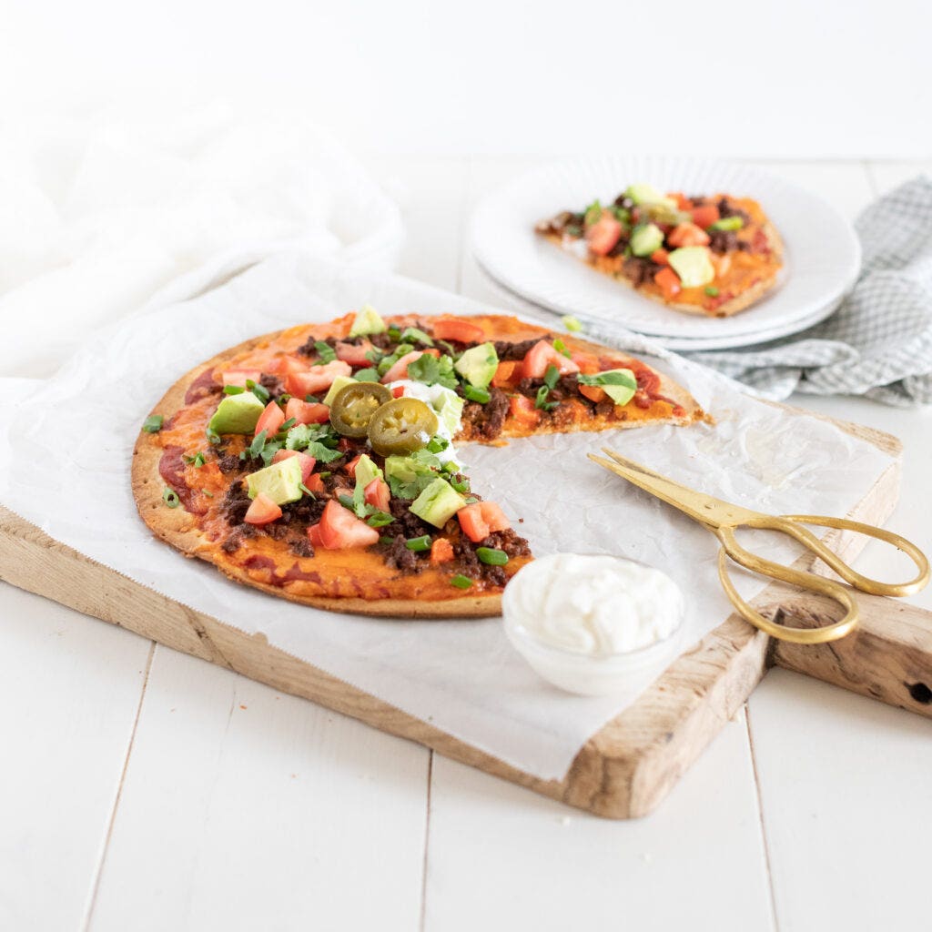 A cutting board with a dinner meal on it which is a Taco Pizza. The pizza is a crust with tomato sauce and topped with taco toppings like  ground meat, tomatoes, avocado, cilantro, jalapeños, and sour cream. A slice is taken out of the pizza and is in the background. This meal is perfect for a summer getaway.