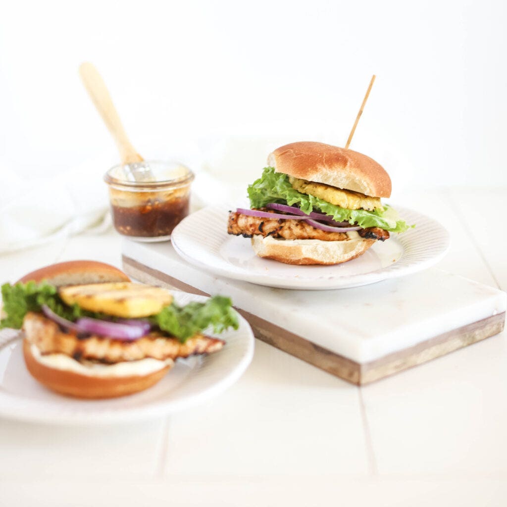 Two plates with a dinner meal on each of them. Each plate has a chicken teriyaki burger on it. The burger is made of a bun, grilled chicken breast with teriyaki sauce, red onion, lettuce, and grilled pineapple. There is a jar of the homemade teriyaki sauce in the background. This meal is perfect for a summer getaway.