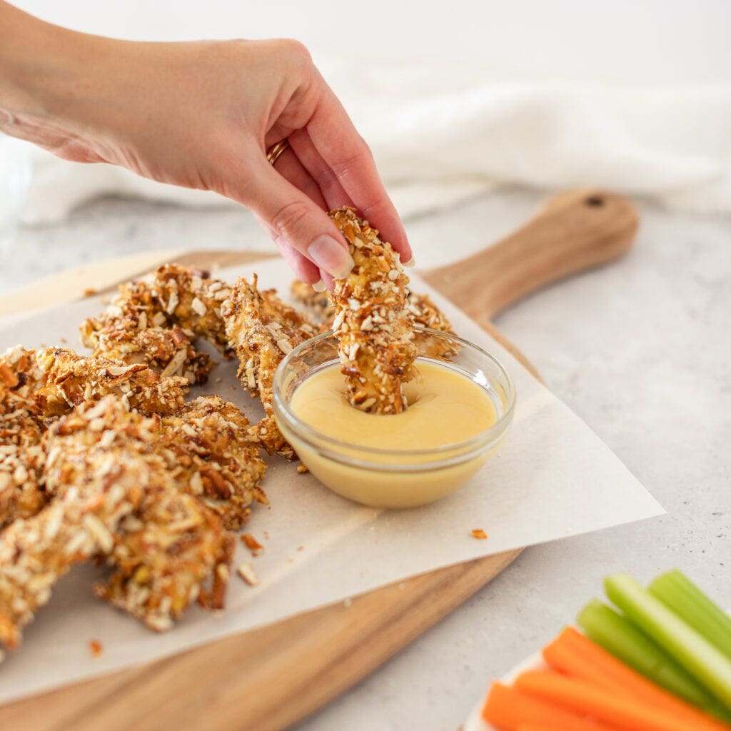 a hand dipping a pretzel crusted chicken finger in sauce