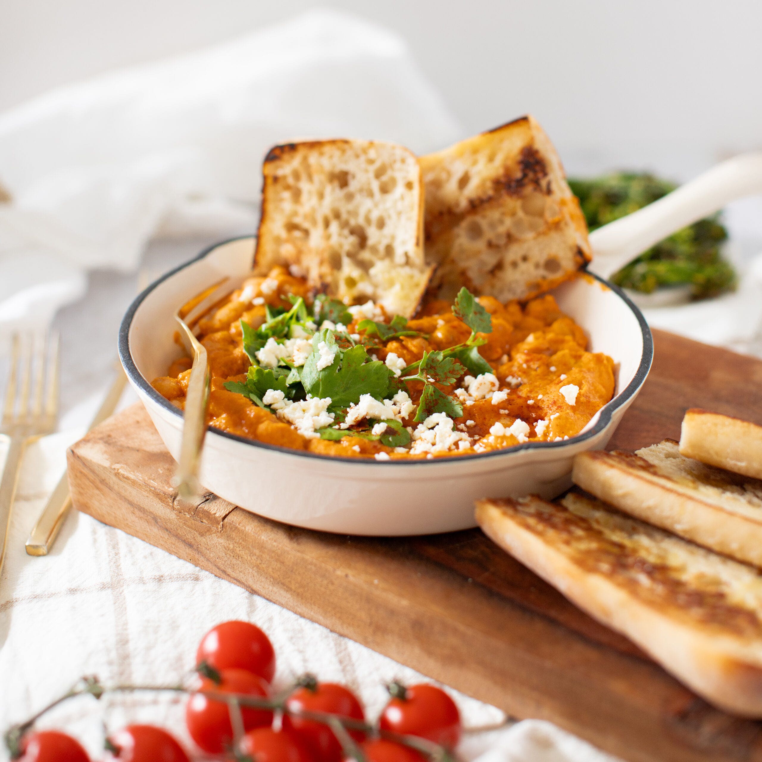 Cannellini beans cooked in a red romesco sauce served in a white cast iron pan with parsley and feta garnish. Grilled ciabatta bread is served on the side.