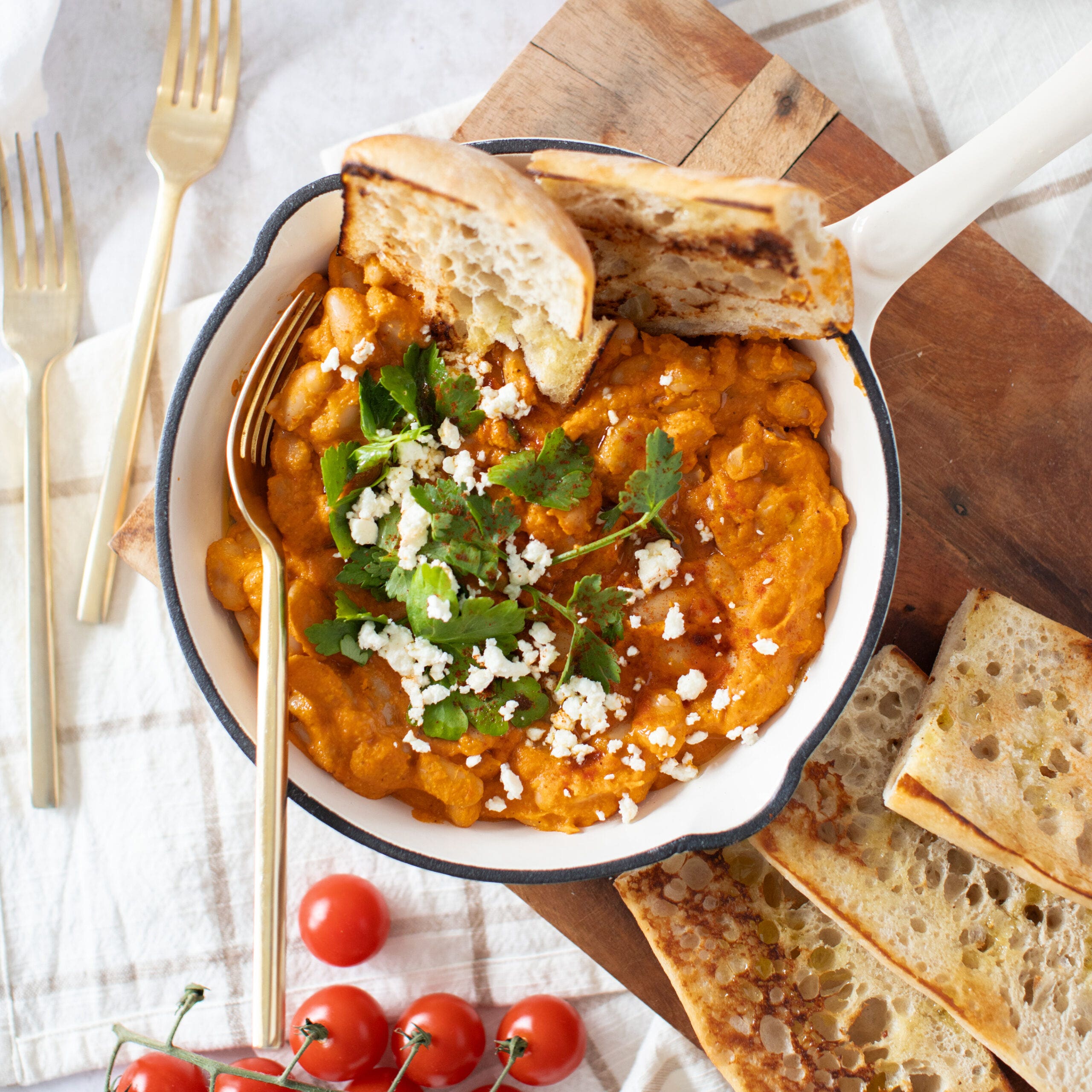 Cannellini beans cooked in a red romesco sauce served in a white cast iron pan with parsley and feta garnish. Grilled ciabatta bread is served on the side.