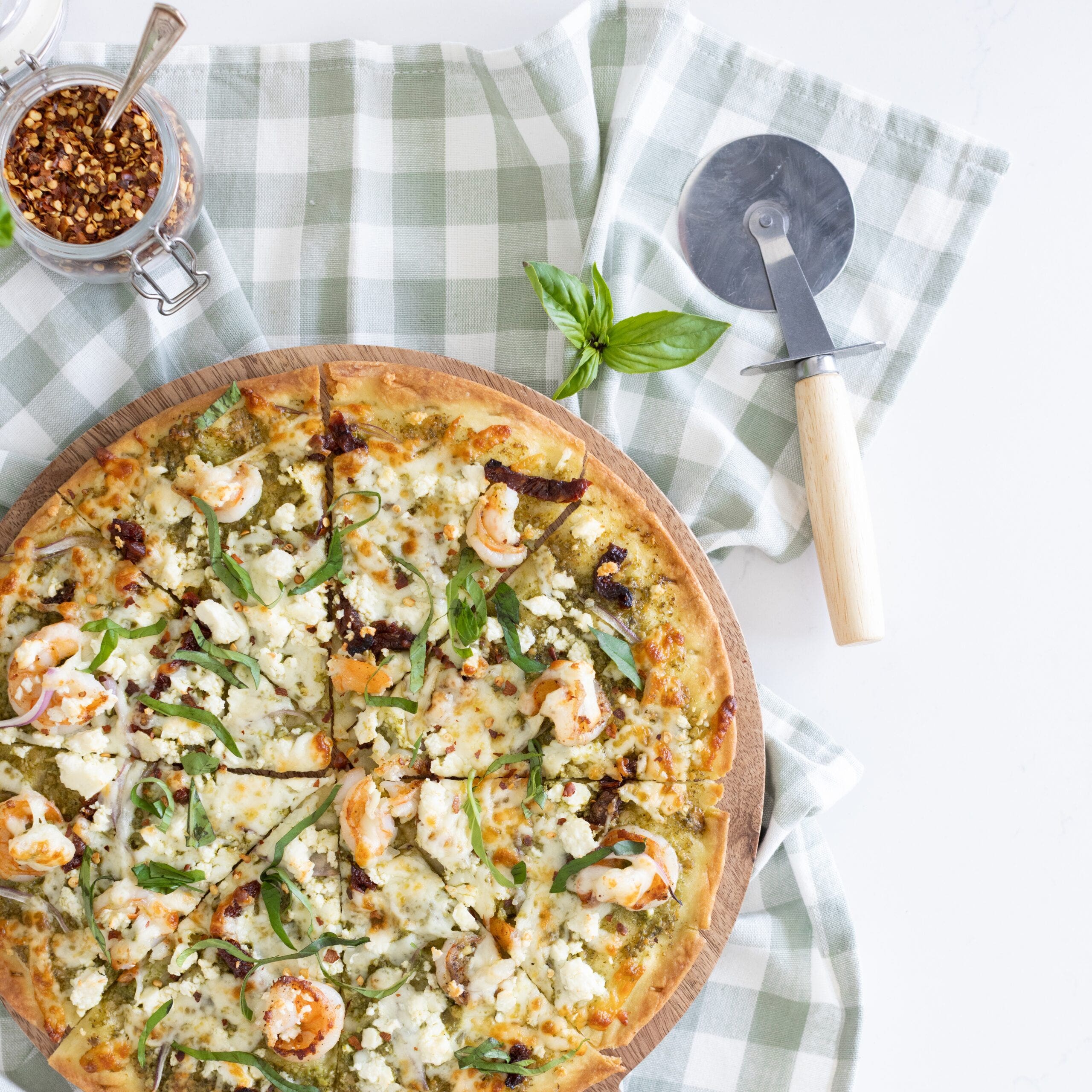 pizza topped with pesto, shrimp, sundried tomatoes and feta with a pizza cutter, red chili flakes and basil around it