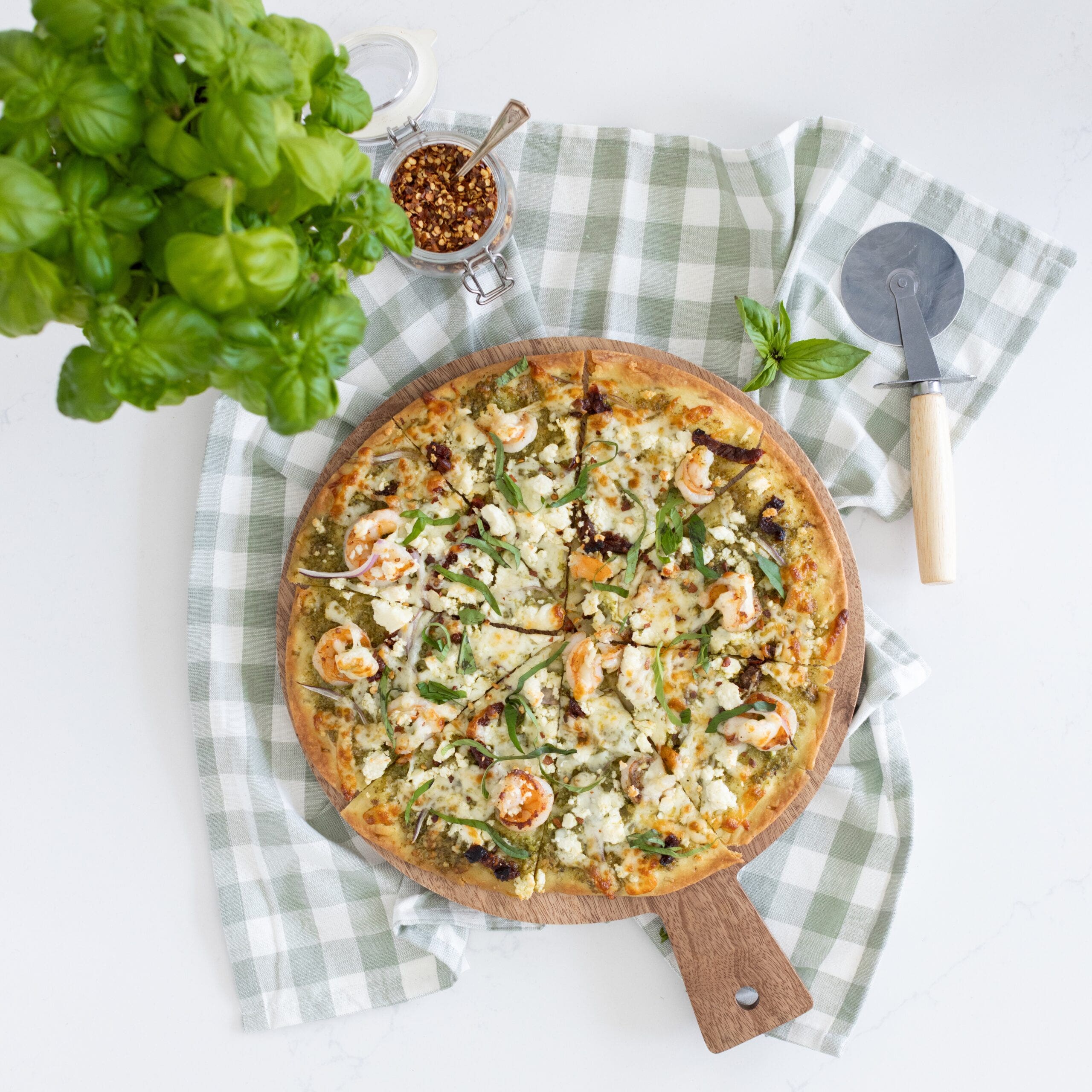 pizza topped with pesto, shrimp, sundried tomatoes and feta with a pizza cutter, red chili flakes and basil around it