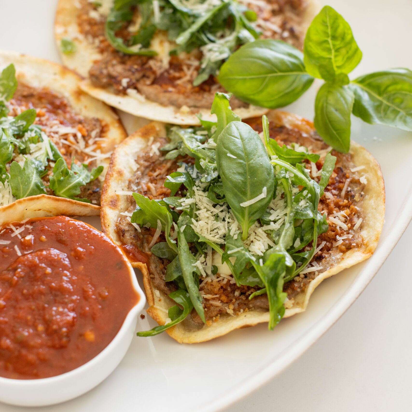 three tacos topped with arugula and basil on a white plate with a side of marinara sauce