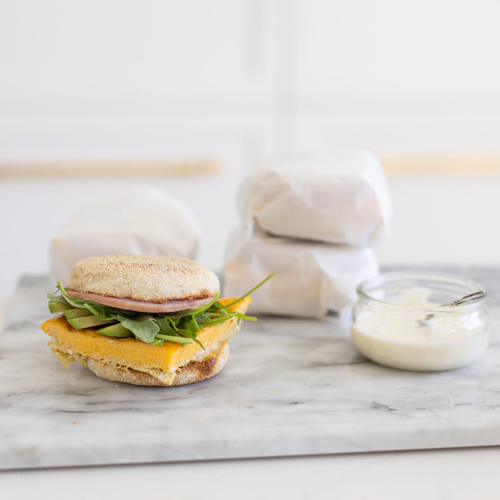 breakfast sandwich on an english muffin with egg, arugula and ham, with parchment wrapped sandwiches in the background