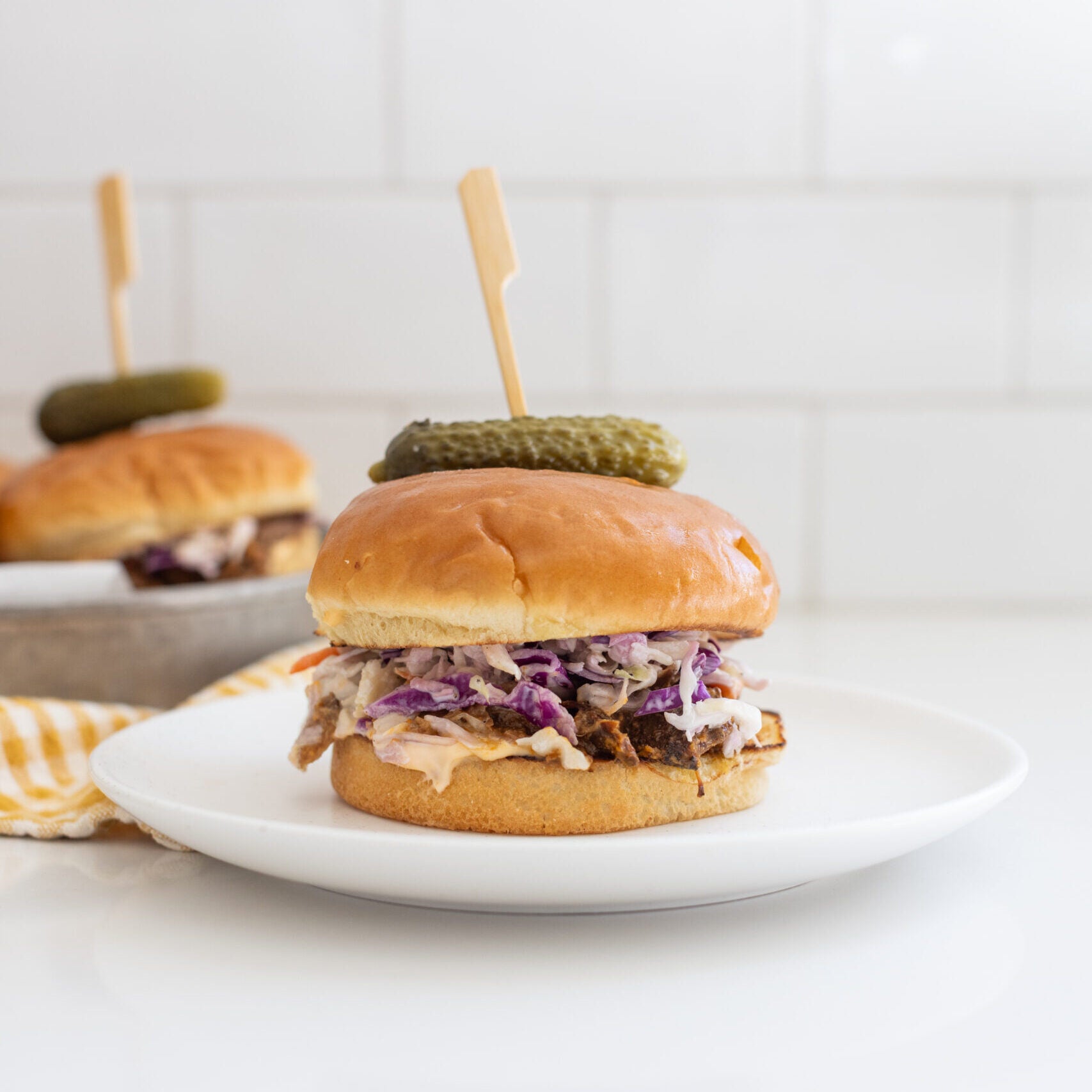 a pulled pork sandwich on a white plate, with a toothpick through it, served with a pickle